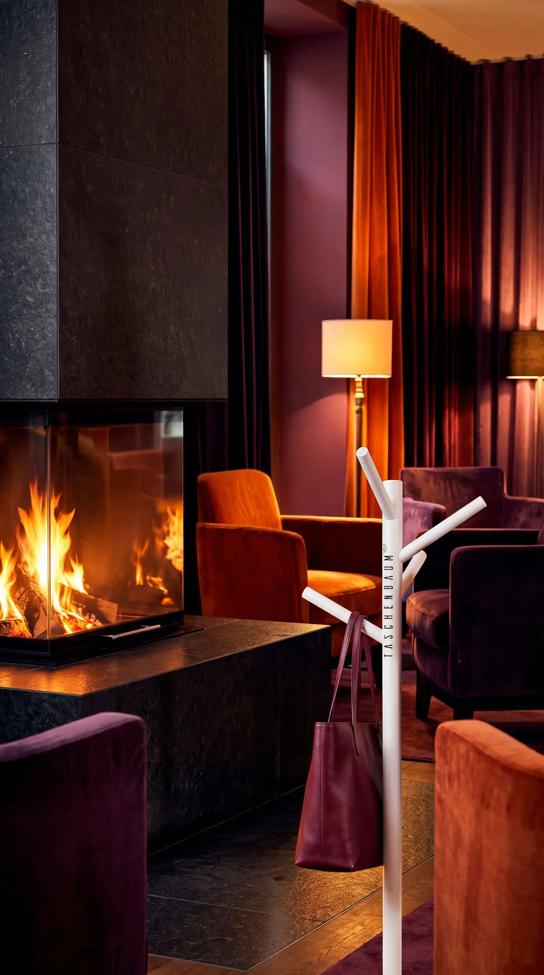 Taschenbaum stehend in einer Hotel Lounge mit Kaminfeuer, er trägt an einem Aste eine Tasche. Das Bild ist im Farbstil violett, orange, beige. Ein Stilbild das in ein Interior-Design Magazin passen würde.